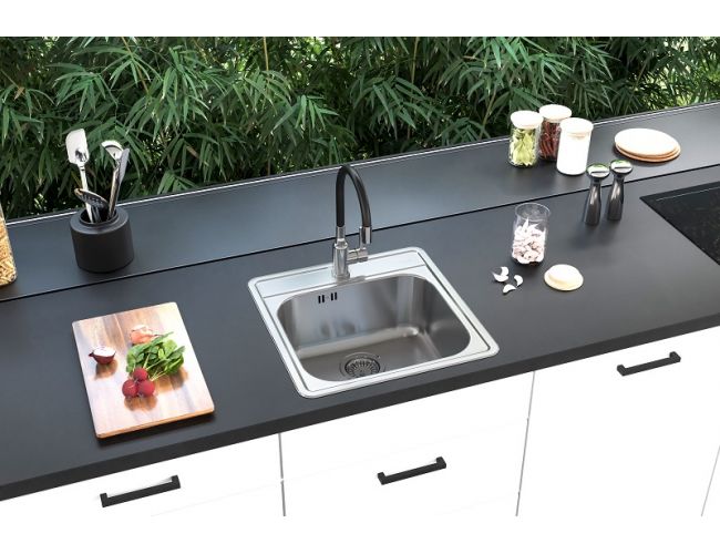 Moderna kuhinja z mat črnim pultom in belimi omaricami, v katerega je vgrajeno kvadratno inox pomivalno korito s črno visoko armaturo; ob njem so deska z zelenjavo, kuhinjski pripomočki in posodice, v ozadju pa steklena stena z razgledom na zelenje.